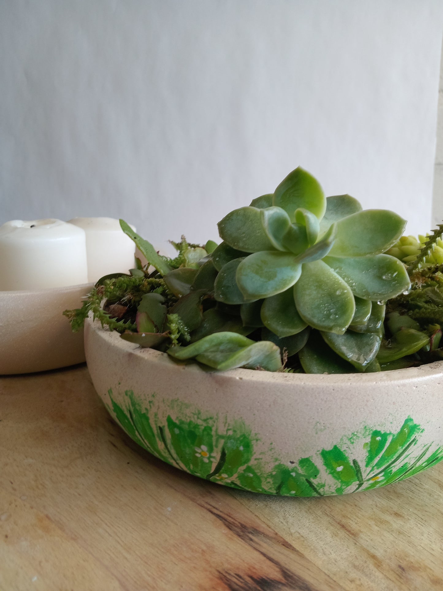 Two beige concrete bowls, one with succulents in them and the other with candles. Both on a wooden surface.