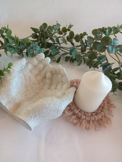 Decorative stone textured giving hands with a candle on a macrame coaster with some greenery around.